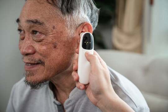Senior Healthcare Checkup with a Caregiver Measuring the Ear Temperature of an Asian Elderly Man in a Detailed Close-Up at Home with Selective Focus