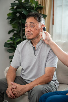 Senior Health Checkup for an Asian Elderly Man Receiving an Ear Temperature Measurement from a Caregiver while Seated at Home in a Vertical Portrait