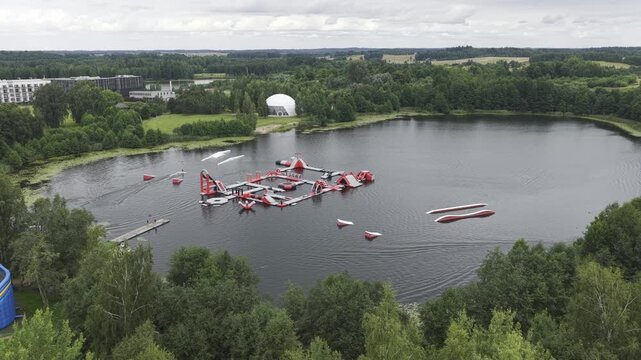 Aerial view of an inflatable water park with red and white obstacles floating on a lake surrounded by lush green forest in Vilnius, Vilnius County, Lithuania.