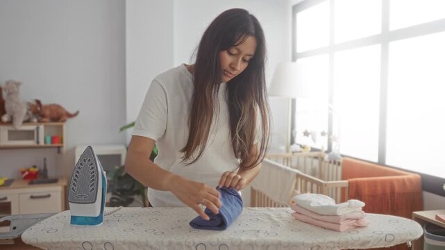 Young latina woman folding baby clothes on an ironing board with iron and stacked garments beside a crib in a nursery building; motherhood care calm.