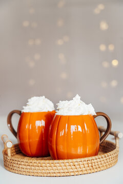 Pumpkin shaped cups of hot drink with whipped cream on woven tray with blurred garland background. Pumpkin latte with spices. Cozy autumn beverage for Thanksgiving or Halloween holidays at home