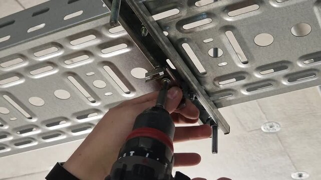 Electrician using a power screwdriver to fix a lamp mounting clip to the bottom of a metal cable tray.