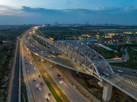 Aerial view of a multi-lane highway featuring a prominent steel truss bridge with evening traffic and surrounding urban development in Gurgaon, Haryana, India.