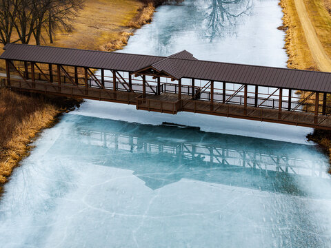 Covered Wooden Bridge Spanning Narrow River with Symmetrical Reflection in Partially Frozen Blue Water and Winter Landscape