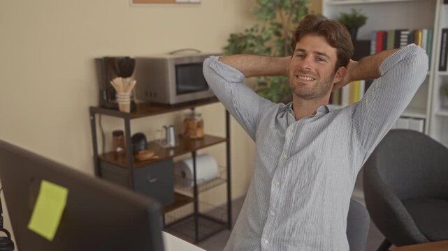 Man sitting at desk, leaning back with hands behind head in office building with computer monitor and bookshelf visible; relaxed productivity.