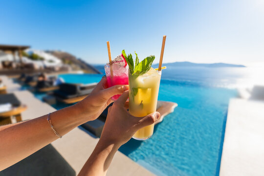 Women's hands hold fresh fruit cocktails with ice. Pool with blue water and sea view in the blurred background. Santorini island, Greece.
