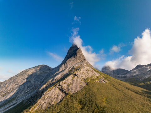Aerial view of Stetind mountain peak with rocky slopes, sparse vegetation, and clouds around the summit under a clear blue sky in Narvik Municipality, Nordland, Norway.