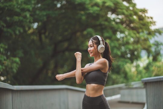 Smiling athletic Asian woman stretching arm shoulder muscle while wearing headphones at city park bridge, fit female runner warming up outdoors before workout for active fitness, health, motivation