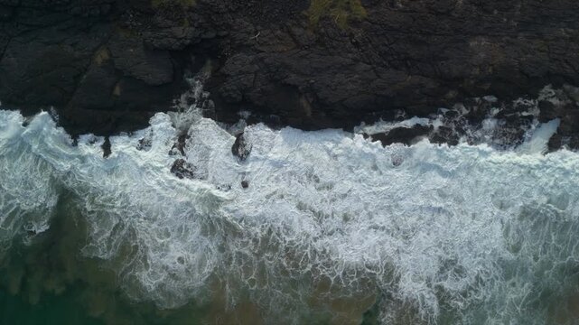Static aerial drone footage looking straight down at ocean waves breaking over the rugged black rocks of South Africa's Wild Coast. Abstract coastal patterns by surf and dramatic shoreline geology