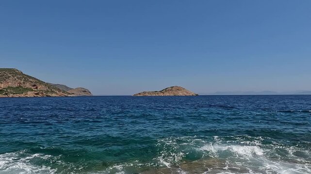 Zoom out footage of a small rocky islet in the Greek sea. Clear blue sky at the background.