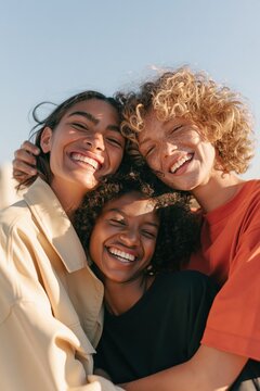 Group of three diverse friends hugging and laughing against clear sky for lifestyle wallpaper and banner use showing candid summer portrait and warm joyful togetherness
