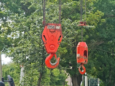 Close-up of two red heavy-duty crane hooks with steel cables suspended against a background of green trees, showing a 25-ton capacity mark.