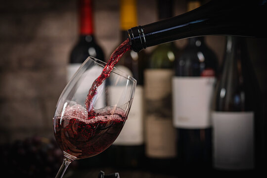 A dark, elegant still life of red wine being poured into a glass with wine bottles and fresh grapes in the background. Perfect for wine-themed compositions