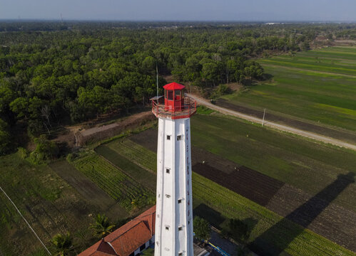Aerial view of a white lighthouse with a red top standing amidst lush green rice fields and agricultural land in Purworejo, Central Java, Indonesia.