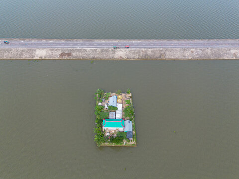 Aerial view of a small homestead on a tiny island surrounded by water, with lush vegetation and a nearby road in Austagram, Dhaka Division, Bangladesh.