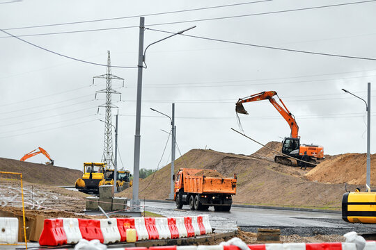 Minsk, Belarus. May 13, 2026. Active road construction site, orange MAZ dump truck carrying soil an orange tracked excavator Hitachi Zaxis working on dirt mound under overcast sky. Road construction