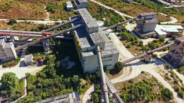 A view of a quarry showing multiple structures, machinery, and conveyor belts operating in a remote area. Dust and vegetation surround the facilities under clear skies.