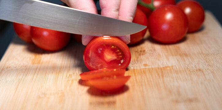 A hand is holding a red tomato on a cutting board and slicing it with a kitchen knife. Freshness, vegetable, contains vitamin C, potassium, vegetarian food, healthy eating, aw food.