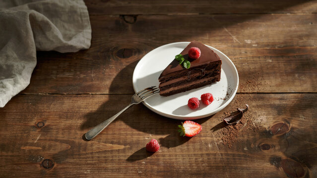 Rich chocolate layer cake slice served with fresh raspberries on a rustic wooden table surface with garnished mint raspberries displays slice decadent