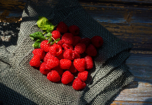 Ripe raspberries, Rubus idaeus, arranged with peppermint leaves, Mentha x piperita, on coarse dark burlap over weathered wood. Dramatic sunlight and shadow emphasize juicy berry texture, summer harves