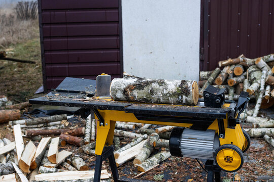 Electric hydraulic log splitter in action outdoors. A large birch log ready for cutting on a yellow machine, surrounded by chopped firewood. Forestry and DIY work. Sawmill