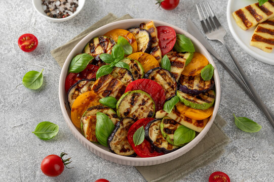 Grilled vegetable salad with zucchini, eggplant, tomatoes, halloumi cheese, basil and balsamic glaze on a plate on a concrete background. Healthy food.