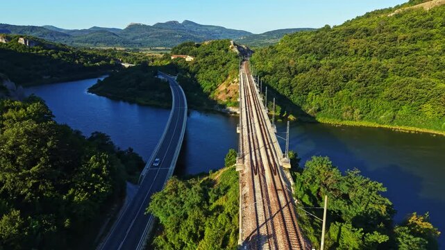Train tracks stretch alongside a river as a highway veers away. Lush green hills rise in the background under a clear sky.