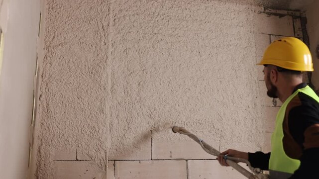 Hard hat, masonry wall, wall coating. On renovation project, construction worker applies cement render to blockwork with spray gun.