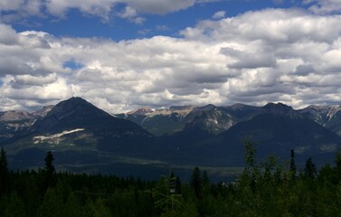 Obraz premium A detailed panoramic view focusing on rugged, dark mountain peaks under a heavily textured blanket of white and grey cumulus clouds with a deep pine forest in the foreground.