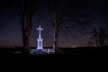 Podświetlony krzyż przydrożny nocą pod rozgwieżdżonym niebem w otoczeniu starych drzew. Symbol wiary, pamięci i tradycji w wiejskim krajobrazie Polski. © Henryk Niestrój