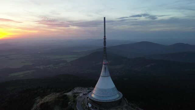 Aerial view of Jested Tower, a unique hyperbolic paraboloid structure atop a mountain, surrounded by rolling hills and forests at dusk in Liberec, Liberecky kraj, Czechia.