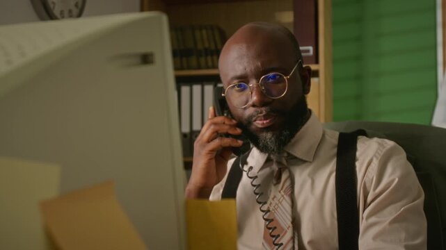 African American man talking on vintage landline telephone while looking at retro computer monitor in office. Footage suits retro business or 1990s corporate nostalgia