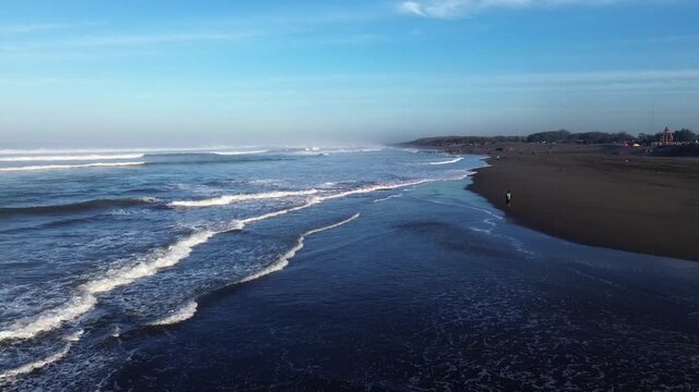 Aerial view of Parangkusumo Beach with dark sand and rolling waves along the coastline in Kecamatan Kretek, Jawa Tengah, Indonesia.
