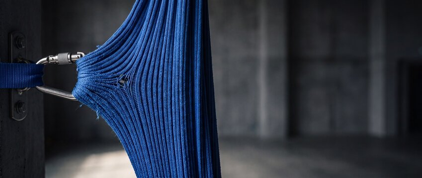 A close up of a strong blue braided rope, showing a slight tear near metal carabiner, secured to a concrete pillar in a dimly lit, industrial background.