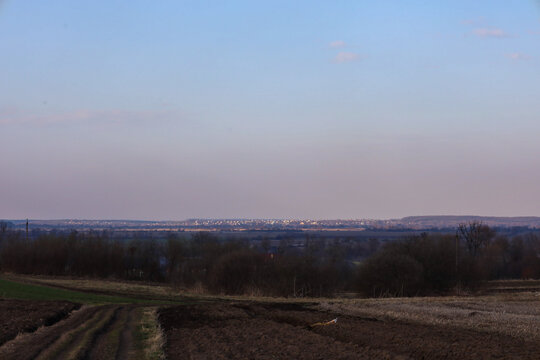 A dirt road winds past freshly plowed fields leading toward a distant village on the horizon under a soft evening sky. The calm rural landscape highlights the quiet atmosphere of the countryside in ea