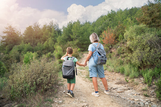 mother and son with backpacks holding hands hiking on forest trail back view
