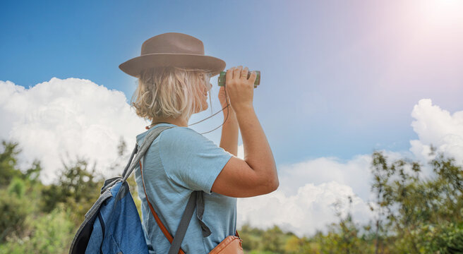 woman tourist with hat and backpack looking through binoculars in nature on sunny summer day