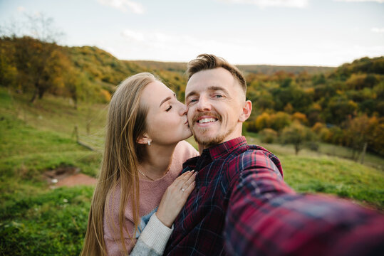 Woman kissing smiling man taking selfie outdoors in autumn mountains. Couple having fun and making self portrait in nature.