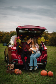 Couple having a cozy picnic with plaid, pumpkins and basket in nature. Woman and man kissing in decorated car trunk holding lantern outdoors in autumn field.
