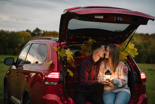 Couple in love hugs in mountains during trip. Woman and man sitting in decorated car trunk holding lantern outdoors in autumn field. Couple having a cozy picnic with plaid and pumpkins in nature.