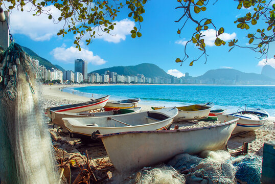 Traditional wooden fishing boats and nets on the white sand of Copacabana Beach, Rio de Janeiro, Brazil, with the city skyline and mountains in the background under a sunny blue sky.