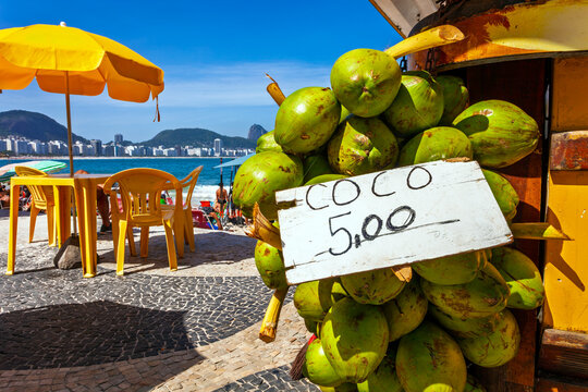 Fresh green coconuts for sale at a Copacabana beach kiosk in Rio de Janeiro, featuring a price sign, the iconic mosaic promenade, and a sunny coast.
