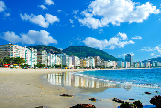 Stunning daytime view of Copacabana beach in Rio de Janeiro, Brazil, featuring the iconic sandy shore, blue ocean waves, vibrant city skyline, and lush mountains under a bright sunny sky.