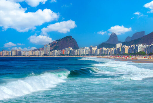 Panoramic view of Copacabana beach in Rio de Janeiro, Brazil. Features crashing ocean waves, the city skyline, and iconic mountains under a bright, sunny blue sky with clouds.