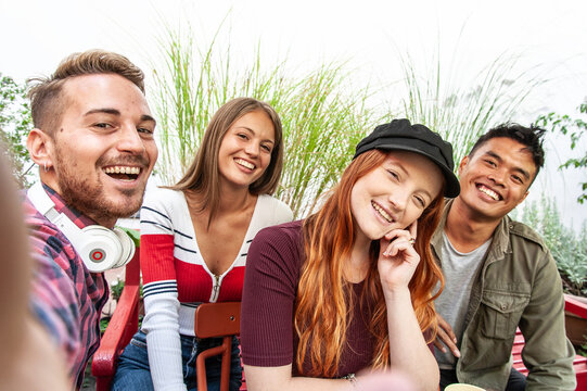 Diverse gen z friends gather for a joyful group selfie while relaxing together on a bright outdoor terrace. Four smiling young adults celebrate friendship, inclusion, and multicultural community.