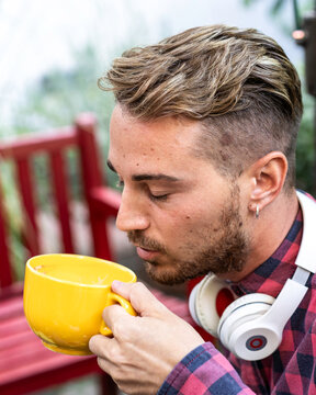 Gen z man savors the aroma of a hot drink from an oversized yellow mug while wearing headphones outdoors. Close-up portrait conveys mindfulness, comfort, music, and relaxation.