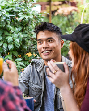 Smiling gen z man chats with friends over hot drinks during a relaxed gathering in a lush outdoor garden. Candid portrait highlights multicultural friendship, conversation, and belonging.