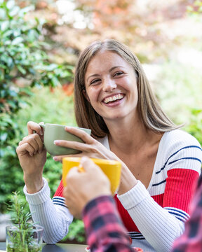 Smiling gen z woman holds a ceramic mug while sharing a cheerful outdoor drink with a friend in a leafy garden. Close-up portrait conveys warmth, connection, and everyday happiness.