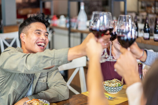 Joyful gen z man raises a glass of red wine during a lively toast with friends at a restaurant. Close-up dining scene highlights celebration, friendship, multicultural connection, and shared joy.