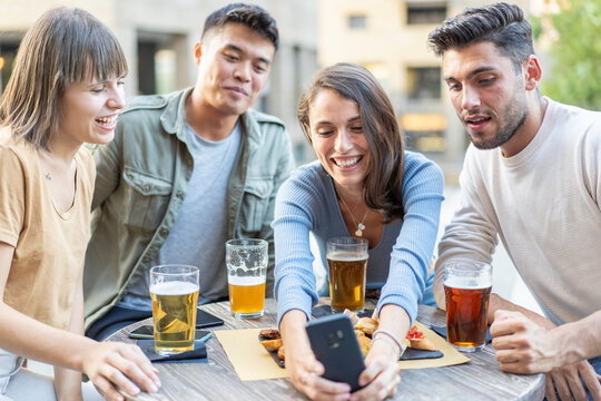 Friends smiling at a smartphone while sharing beers at an outdoor cafe. Multiethnic millennials and gen z gather around the table for a digital memory. Social media concept